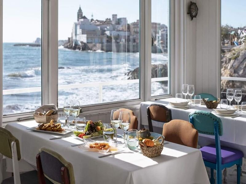Mesa de restaurante junto al mar en Sitges, con la iglesia de Sant Bartomeu i Santa Tecla al fondo