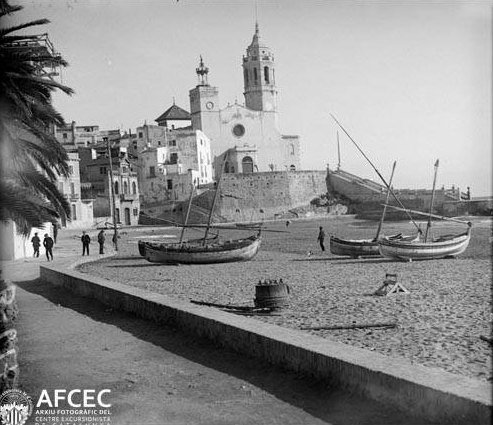 Barcas varadas en la arena de la playa de la Ribera de Sitges, con la iglesia de Sant Bartomeu i Santa Tecla al fondo, hacia 1920–1930