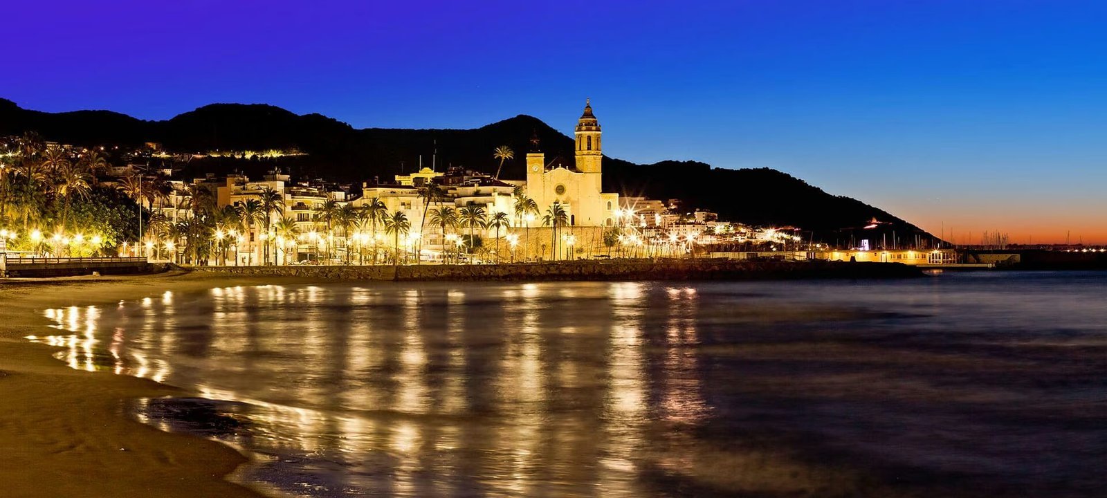 Sitges de nit: el passeig il·luminat i l’església de Sant Bartomeu i Santa Tecla reflectits a la platja